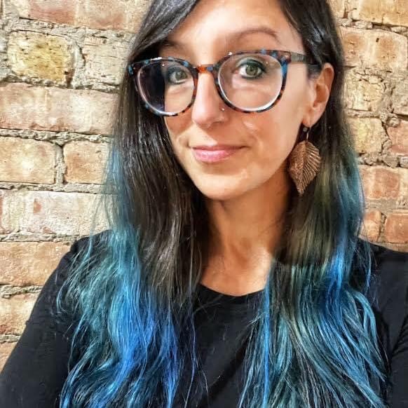 Woman with long dark hair with blue highlights wearing glasses and a black top, smiling in front of a brick wall